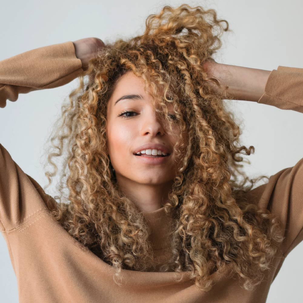 Une personne aux cheveux bouclés blonds porte un pull beige et sourit devant un fond neutre.