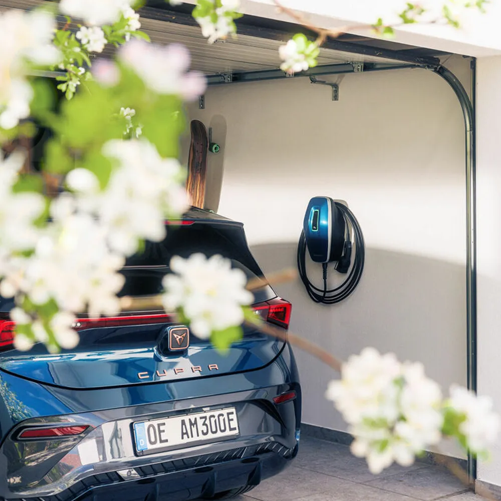 Une voiture bleue est garée dans un garage avec une borne de recharge murale, entourée de branches fleuries au premier plan.