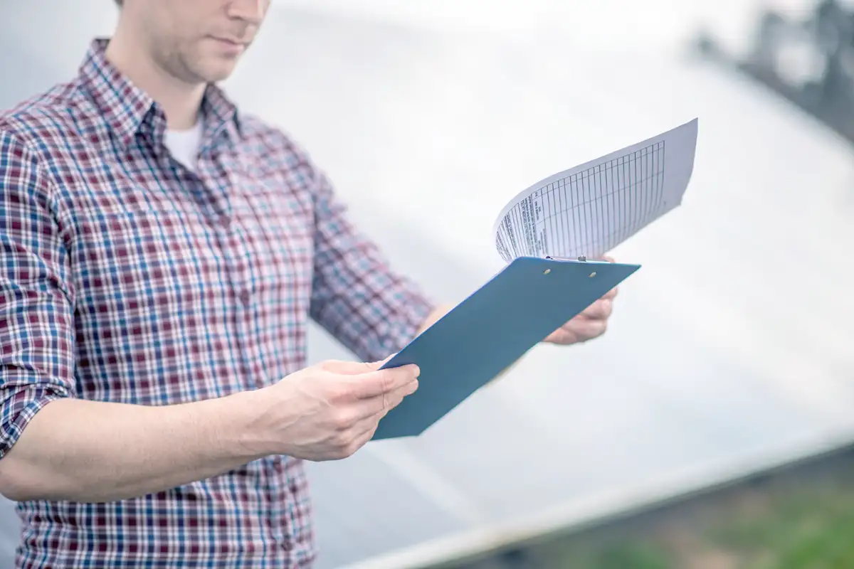 Un homme en chemise à carreaux tient un classeur bleu avec des documents devant des panneaux solaires.