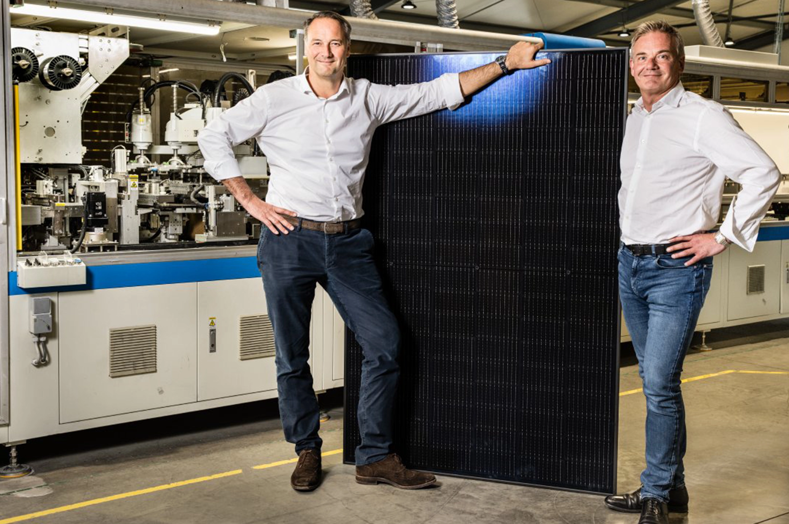 Deux hommes en chemise blanche posent avec un panneau solaire noir dans une usine aux machines industrielles.