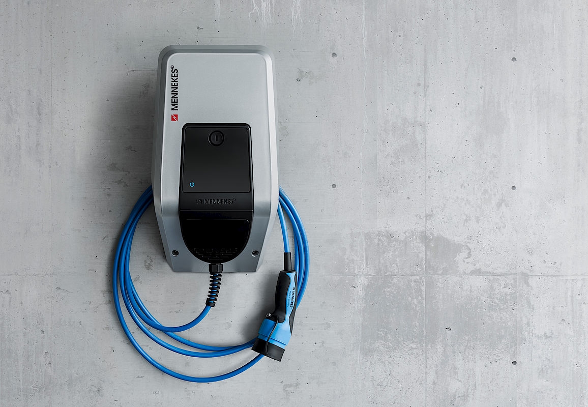 Borne de recharge grise avec câble bleu fixée à un mur en béton, portant le logo Mennekes.