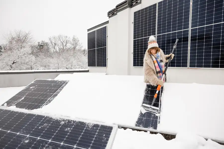 Une personne déneige des panneaux solaires sur un toit enneigé, vêtue d'un manteau beige et d'un bonnet à pompon.