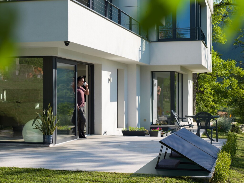 Un homme boit un café devant une maison moderne avec des panneaux solaires, des chaises noires et des plantes en pot.