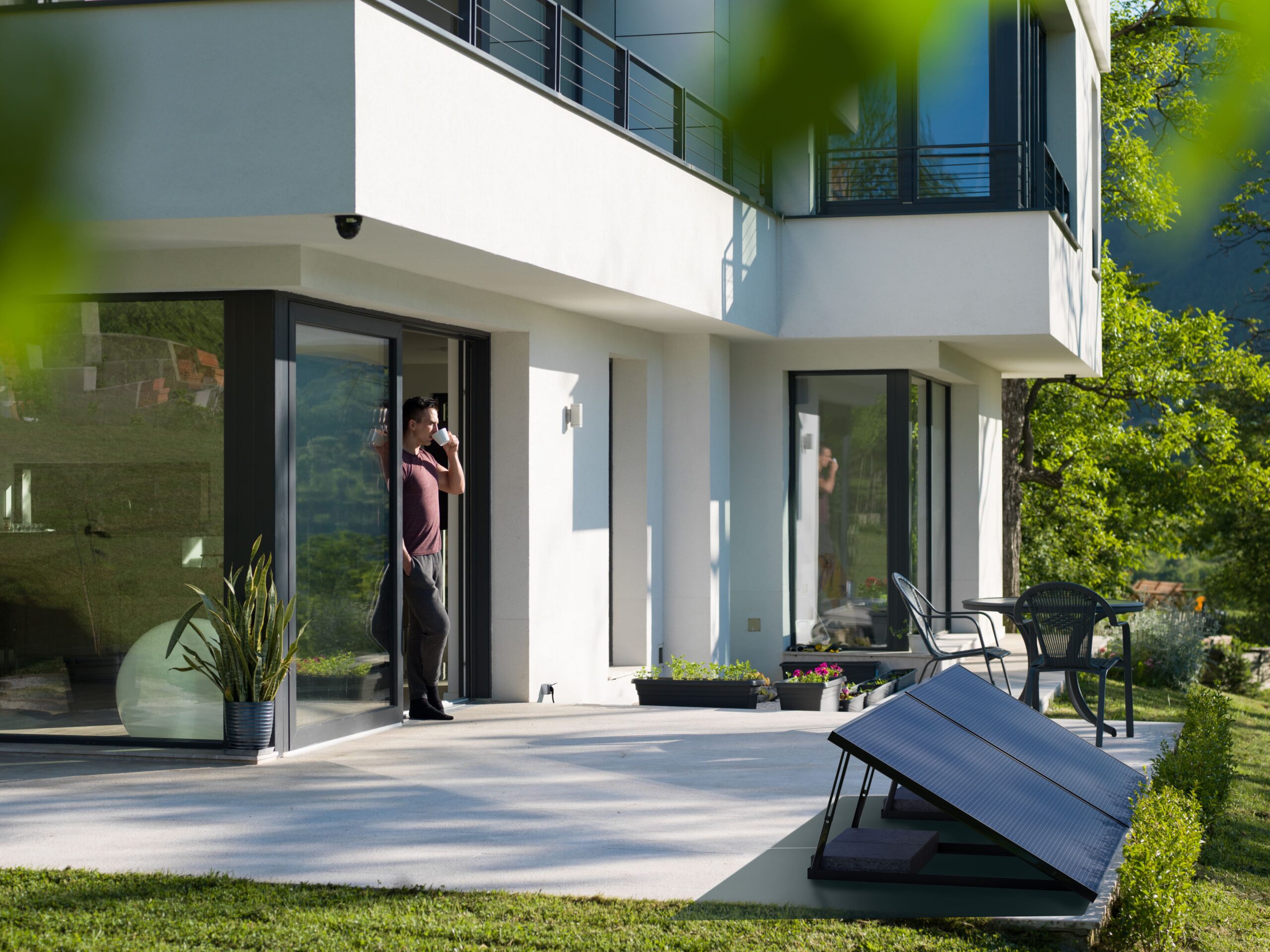 Un homme boit un café devant une maison moderne avec des panneaux solaires, des chaises noires et des plantes en pot.