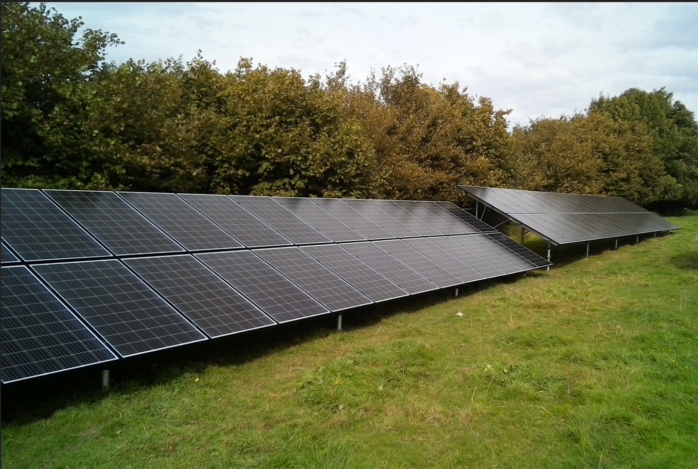 Panneaux solaires noirs installés sur une pelouse verte, avec des arbres en arrière-plan sous un ciel partiellement nuageux.